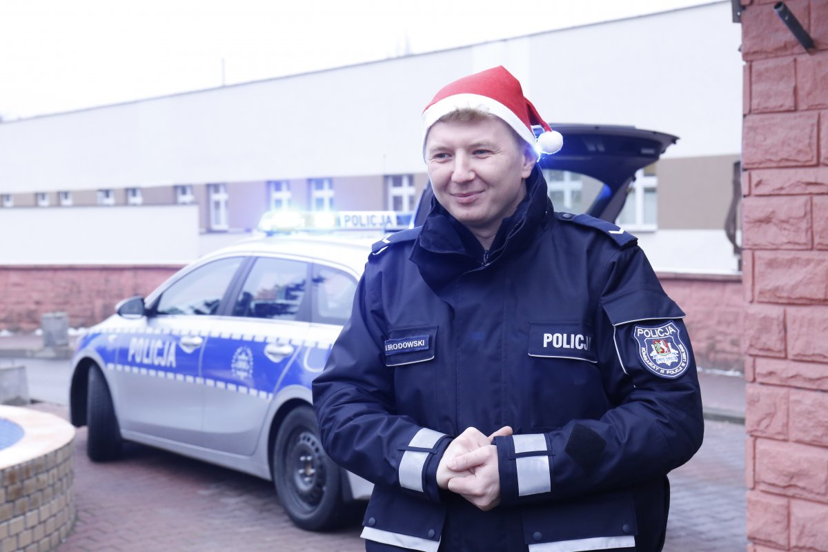 Policjan ubrany w mundur z założoną na głowę czapką Mikołaja. W drugim planie jest oznakowany radiowóz. 