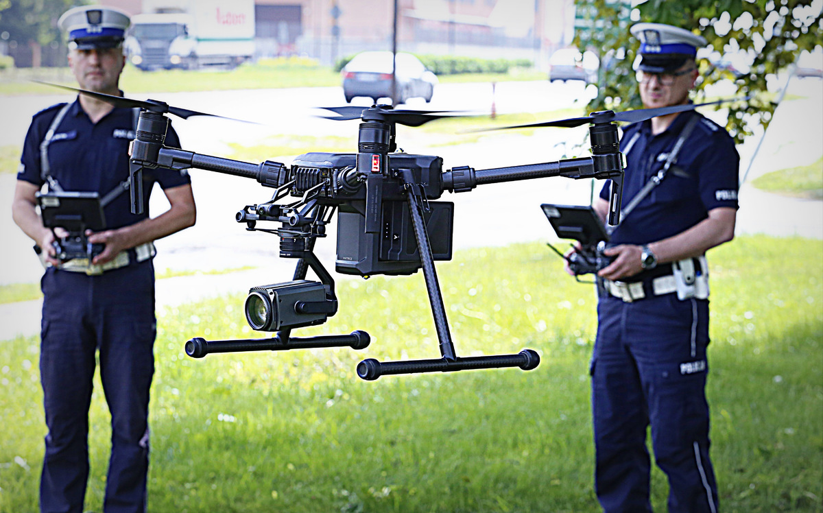Zdjęcie przedstawia w pierwszym planie drona unoszącego się w powietrzu i sterujących nim policjantów w drugim planie.