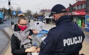 Policjant przekazuje maseczkę kobiecie, która trzyma na rękach dziecko.