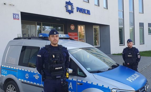 policjanci przy radiowozie, w tle widać budynek komisariatu policji