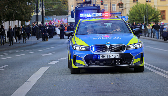 Oznakowany radiowóz w pierwszym planie zdjęcia. W głębi policjanci zabezpieczający marsz równości.